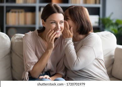 Middle aged mother whispering in smiling adult daughter ear, gossiping, sharing secret, happy beautiful woman hearing good news from mature mum, sitting together on couch, trusted relations