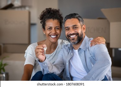 Middle aged multiethnic couple embracing and holding house keys with carboard boxes behind them. Portrait of indian man and african woman showing house keys while looking at camera after relocation.