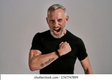 Middle Aged Muscular Man In Black T Shirt Shouting At Camera, Beating His Chest While Posing In Studio Over Grey Background. Horizontal Shot