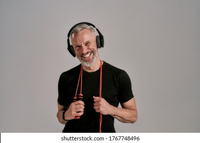 Middle aged muscular man in black t shirt and headphones smiling at camera holding jump rope, posing in studio over grey background. Sport, healthy lifestyle concept. Front view. Horizontal shot - Powered by Shutterstock