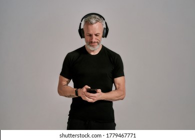 Middle Aged Muscular Man In Black T Shirt And Headphones Using Smartphone While Posing In Studio Over Grey Background. Front View. Horizontal Shot