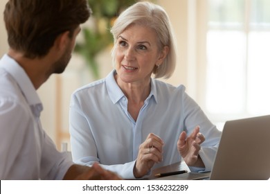 Middle aged old businesswoman manager mentor speaking to businessman client offering insurance banking services deal teaching intern or convincing customer at business office meeting sit at table.