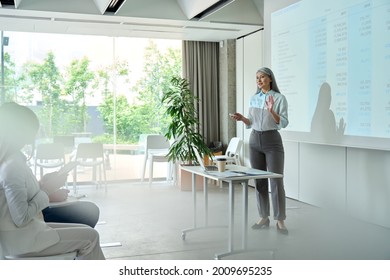 Middle aged older happy Asian businesswoman coach mentor speaker executive presenting teaching business seminar to students team standing in board meeting room in modern office conference workshop. - Powered by Shutterstock