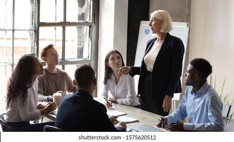 Middle aged professional coach educating focused mixed race managers at office. Concentrated young multiracial coworkers listening to older speaker, presenting project ideas at brainstorming meeting.