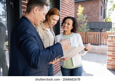 Middle Aged Realtor Holding Folder Near Smiling Interracial Lesbian Couple