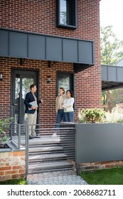 Middle Aged Realtor Pointing At House With Brick Wall Near Interracial Lesbian Couple