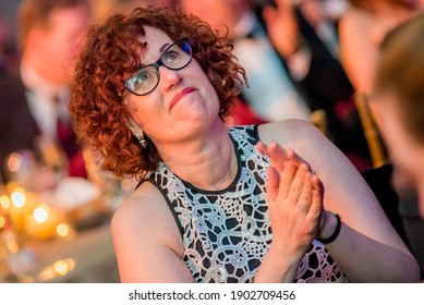 A Middle Aged Red Curly Haired White Woman With Dark Glasses Looks Up In Wander During A Black Tie Event. She Is Smiling And Clapping Looking At The Stage.