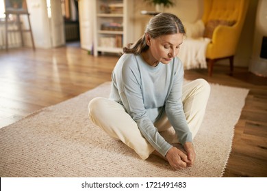 Middle Aged Senior Caucasian Woman Sitting On Carpet In Living Room Doing Butterfly Pose Or Baddha Konasana To Strengthen And Open Hips, Holding Hands On Her Feet. Yoga, Flexibility And Vitality