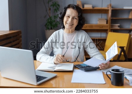Middle aged senior woman sit with laptop and paper document. Smiling older mature lady reading paper bill pay online at home managing bank finances calculating taxes planning loan debt pension payment