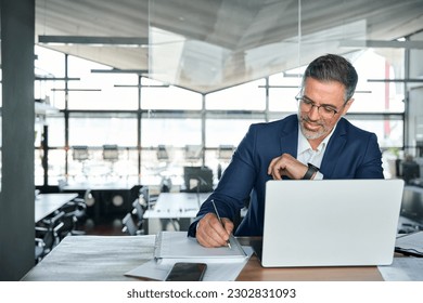 Middle aged smiling professional business man global company executive ceo manager or lawyer wearing suit sitting at desk in modern office working on laptop computer and writing notes, copy space.