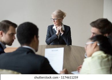 Middle aged stressed woman sitting at the desk in office at meeting. Young multiracial workers team sitting their backs to camera. Passing interview, hiring, recruiting and age discrimination concept