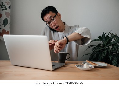 Middle Aged Surprised Asian Man Looking At Watch Talking By Smartphone And Working With Laptop While Eating Breakfast In Kitchen.