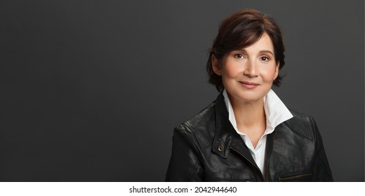 Middle aged well looking woman portrait. Woman wearing white shirt and black leather jacket against dark solid background. Studio female portrait. Billdoard size. - Shutterstock ID 2042944640