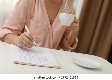 Middle aged woman with cup of drink solving sudoku puzzle at white table indoors, closeup