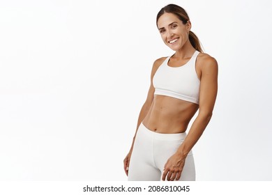 Middle aged woman doing fitness workout, standing in activewear with abs and muscles, smiling happy, standing over white background