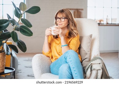 Middle aged woman drinking her tea and daydreaming while sitting in armchair at home.  - Powered by Shutterstock