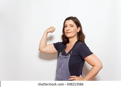 Middle aged woman in dungarees with one hand on her hip and flexing the biceps muscles of the other arm, white background