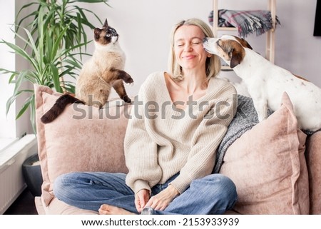 Middle aged woman enjoys spending time at home with her pets. Dog licks owner's cheek with his tongue, cat sitting on couch