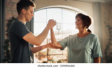 Middle Aged Woman Exercising At Home With Personal Trainer. Elderly Female Strengthening Arm Muscles With Dumbbell Workout. Young Man Supporting And Guiding Her Through The Training.