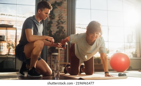 Middle Aged Woman Exercising At Home With Personal Trainer. Elderly Female Strengthening Core And Arm Muscles With Dumbbell Workout. Young Athlete Supporting And Guiding Her Through The Training.