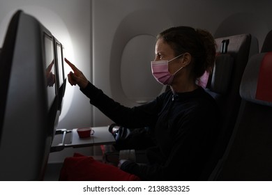 Middle Aged Woman With Face Covering Using Touchscreen Entertainment System On Airplane Seat. Female Passenger On Plane During Pandemic