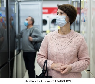 Middle Aged Woman In Face Mask Standing In Appliance Store And Choosing Refrigerator.