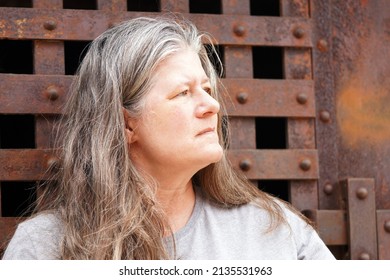 Middle Aged Woman With Gray And Brown Long Hair Looks Right Against An Old Iron Gate With Rivets                               