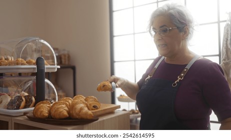 Middle aged woman with grey hair and glasses serving pastries in a bakery shop with shelves of assorted baked goods. - Powered by Shutterstock