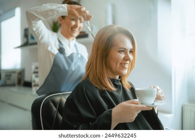 middle aged woman hair salon employee in modern hair studio cutting hair with scissors and relaxed client with cup of tea. - Powered by Shutterstock