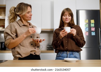 Middle aged woman and her adult daughter drink coffee and gossip in the kitchen. Teenage girl visiting mom - Powered by Shutterstock