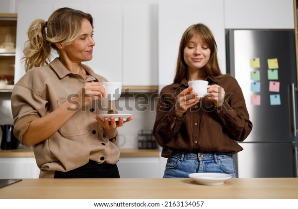 Middle aged woman and\
her adult daughter drink coffee and gossip in the kitchen. Teenage\
girl visiting mom