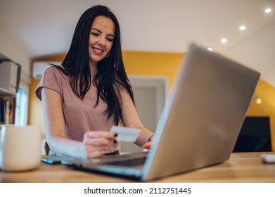 Middle Aged Woman Holding Credit Card And Using Laptop While In The Kitchen At Home.