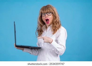 middle aged woman holding laptop with surprised gesture on blue background - Powered by Shutterstock