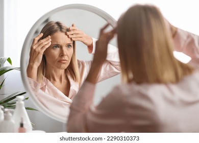 Middle Aged Woman Looking In Mirror And Touching Wrinkles On Her Face, Upset Beautiful Mature Female Examining Fine Lines On Forehead, Suffering Skin Aging, Selective Focus On Reflection, Closeup - Shutterstock ID 2290310373