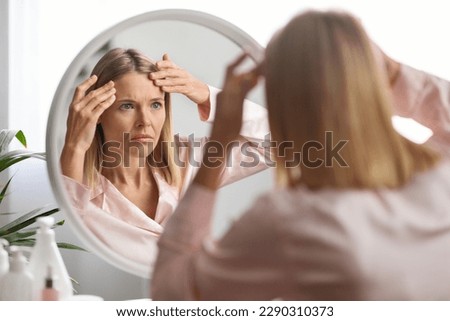Middle Aged Woman Looking In Mirror And Touching Wrinkles On Her Face, Upset Beautiful Mature Female Examining Fine Lines On Forehead, Suffering Skin Aging, Selective Focus On Reflection, Closeup