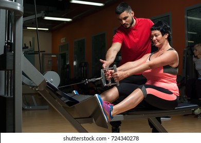 Middle Aged Woman On Exercising Machine And Her Personal Coach. Young Male Trainer Assisting Female Client In Gym. Healthy Lifestyle, Fitness And Sports Concept.