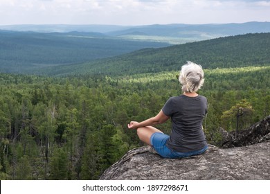 A middle aged woman practices yoga and meditation on a mountain top with a stunning view of a forested, hilly valley. - Powered by Shutterstock