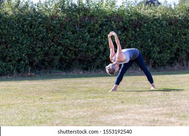 A Middle Aged Woman Practicing Yoga Barefoot Outside In A Grassy Park. She Is Wearing A Grey And Blue Vest And Black Leggings. The Style Of Yoga She Is Doing Is Hatha Yoga As Well As Some Reiki