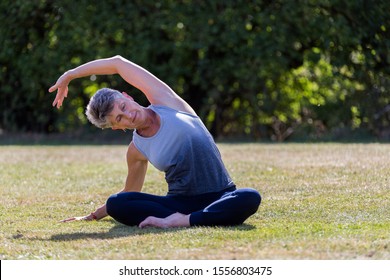 A Middle Aged Woman Practicing Yoga Barefoot Outside In A Grassy Park. She Is Wearing A Grey And Blue Vest And Black Leggings. The Style Of Yoga She Is Doing Is Hatha Yoga As Well As Some Reiki