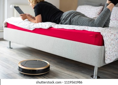 A middle aged woman is reading on her digital tablet on her bed while her vaccum cleaning robot is cleaning the wooden floor at home. Household, leisure and technology concept
