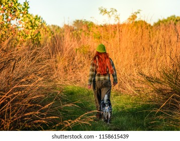 Middle Aged Woman With Red Hair Walking On The Trail With A Dog Behind Her In The Fall; Tall Grasses On Both Sides; Missouri, Midwest