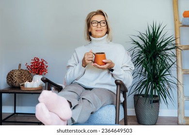 Middle aged woman relaxing with cup of hot drink in scandy style hygge interior home with fall mood interior decor. Lady dreaming, enjoy calm mood without stress, wellbeing alone. Cozy autumn at home.