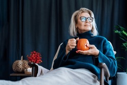 Middle Aged Woman Relaxing With Pumpkin Shaped Cup Of Hot Drink In Scandy Style Hygge Interior Home With Fall Mood Decor. Lady Dreaming, Enjoy Calm Mood Without Stress, Well Being Alone. Cozy Autumn