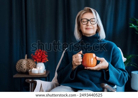 Middle aged woman relaxing with pumpkin shaped cup of hot drink in scandy style hygge interior home with fall mood decor. Lady dreaming, enjoy calm mood without stress, well being alone. Cozy autumn.