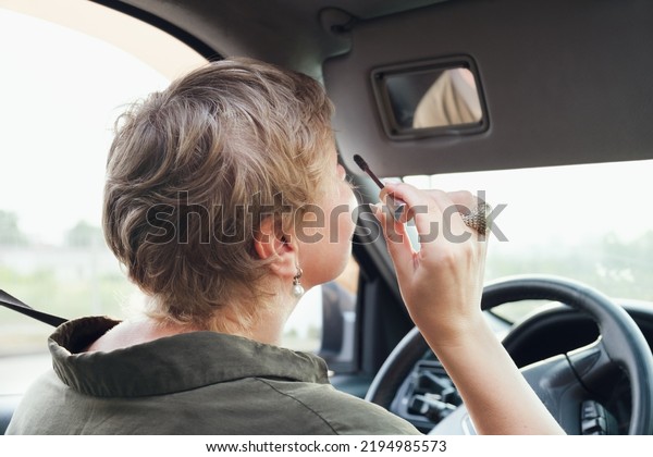 Middle aged woman\
with short hair driving a car tint eyelashes with mascara. The\
woman driving making make-up looking in the Decorative makeup\
cosmetic mirror for sun\
visor