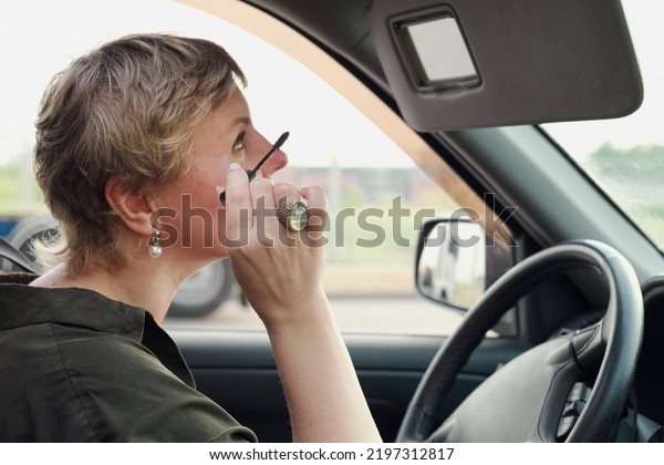 Middle aged woman
with short hair driving a car tint eyelashes with mascara. The
woman driving making make-up looking in the Decorative makeup
cosmetic mirror for sun
visor