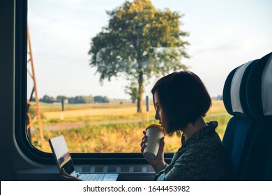 A middle aged woman sitting by the window of a commuter train with a laptop and coffee in a reusable bamboo cup in hand. The concept of remote work, business trips, freelancer, Tourist solo travel