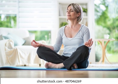 Middle Aged Woman Sitting In Lotus Position On A Carpet In His Living Room. Her Eyes Are Closed. She Is In The Foreground, The Focus Is On It. In The Background, The Garden Is Blur