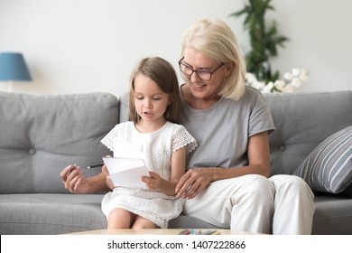 Middle Aged Woman Sitting On Couch At Home With Little Girl. Loving Grandma Teaching Adorable Preschool Grandchild Reading Or Counting Numbers Or To Draw. Education And Development Of Child Concept