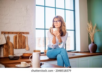 Middle aged woman sitting on the kitchen’s counter and drinking her morning tea while daydreaming.  - Powered by Shutterstock